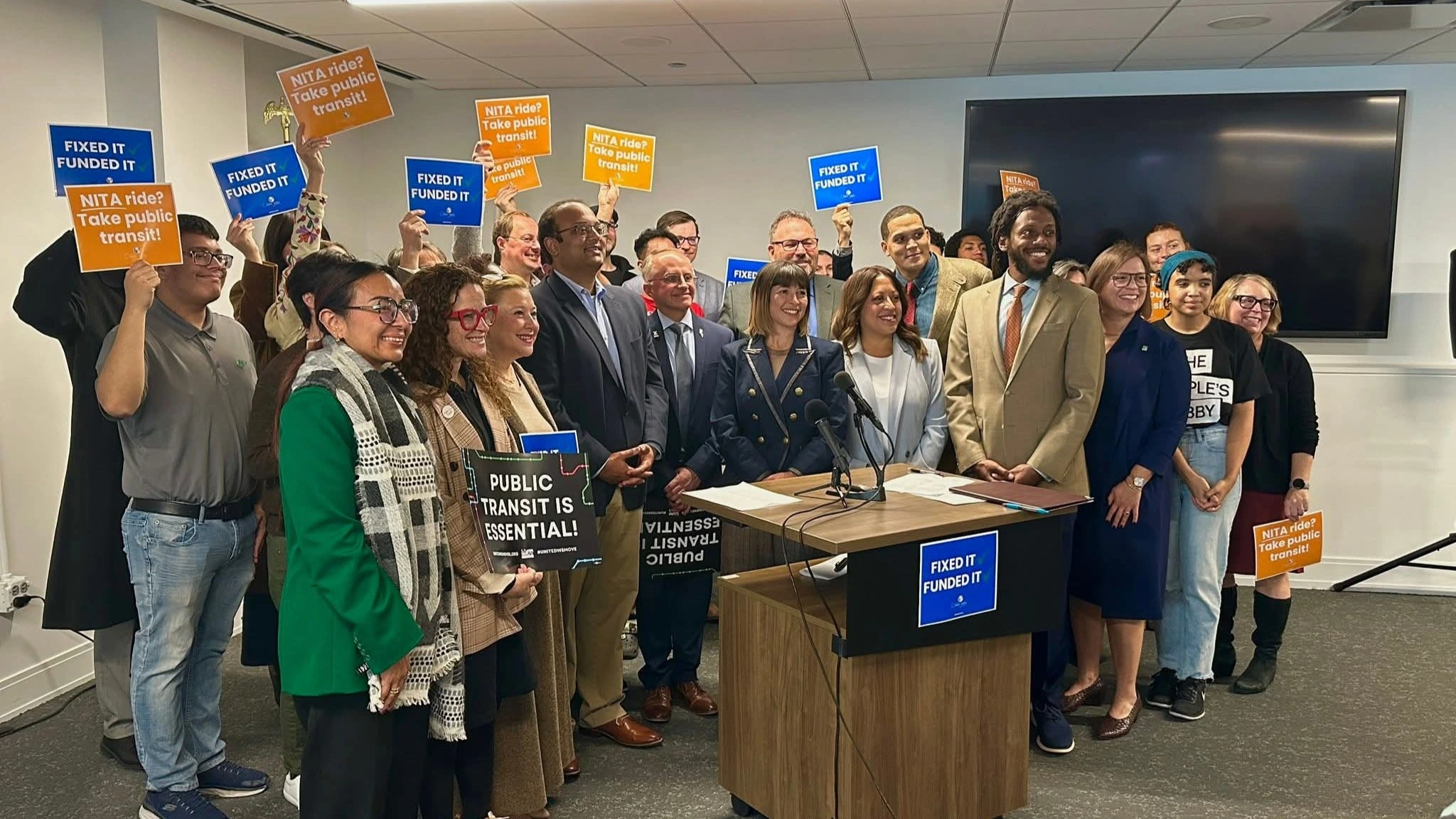 A group of about thirty people smiling and holding up pro-public transit signs in a conference room.